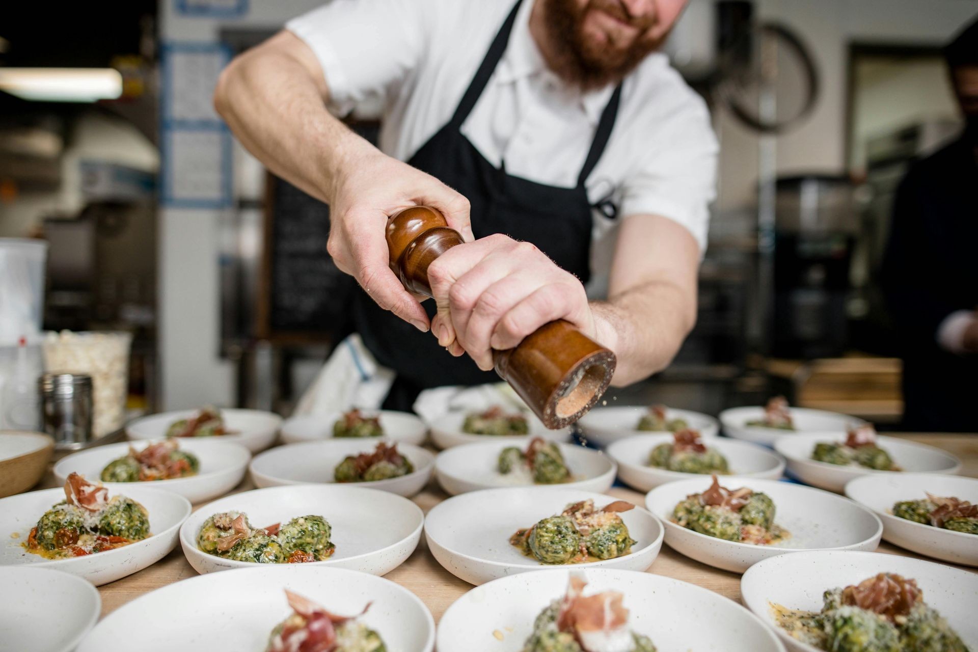 Chef in an apron seasoning multiple plates of food with a pepper grinder in a busy kitchen.